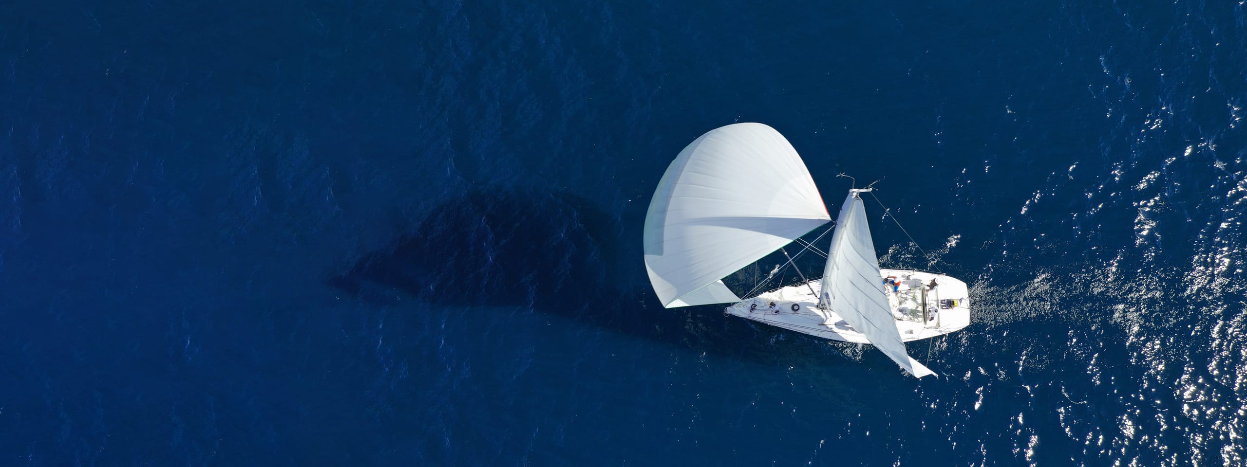 A sailboat under a full spinnaker seen from directly above, its sail shadow visible beneath the deep blue water